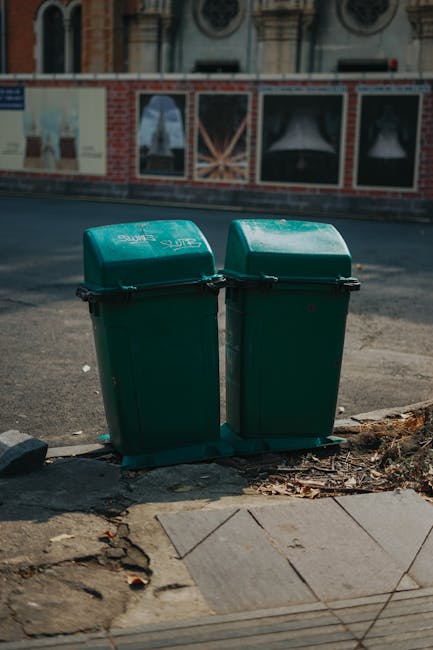Two green wheelie bins with hinged lids are positioned side by side on a pavement near the edge of a curb. The bins are made of plastic with a smooth, slightly reflective surface, and are secured with black clips. They are placed on a small concrete base, with some scattered leaves and dirt around the base. Behind them, there is a city street with an asphalt road, which appears clean and well-maintained. In the background, a brick building with decorative stone or concrete elements, including arched windows and circular ornamentation, is visible, along with a large billboard or poster panel mounted on the building's exterior wall. The scene is illuminated by daylight, with a neutral, slightly overcast atmosphere. The presence of the waste bins near the curb suggests they are set out for collection, aligning with private or alternative waste handling practices often managed by companies such as House Clearance Finsbury Park, rather than local authority collection services.