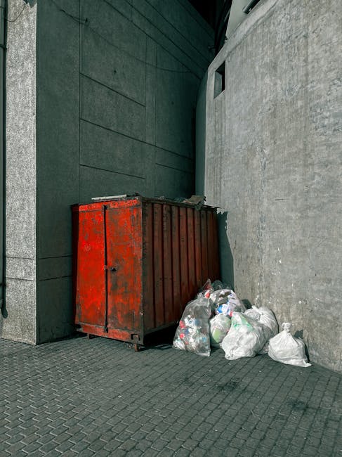 A large, red metal waste skip with visible rust and chipped paint is positioned in the corner between a dark green paneled wall and a light grey concrete wall. The skip, used for rubbish removal, is set on a paved surface with small, dark grey interlocking bricks. Several white plastic bags filled with mixed waste, including recycling materials such as plastic bottles and packaging, are strewn next to the skip, indicating recently collected or awaiting disposal. The scene appears to be outdoors, possibly in an alley or service area adjacent to a building, with natural daylight casting soft shadows. The overall setting suggests a site for independent disposal or private waste collection, typical of rubbish removal services like those offered by House Clearance Finsbury Park. The environment is utilitarian and functional, emphasizing waste sorting and removal without additional decorative elements.