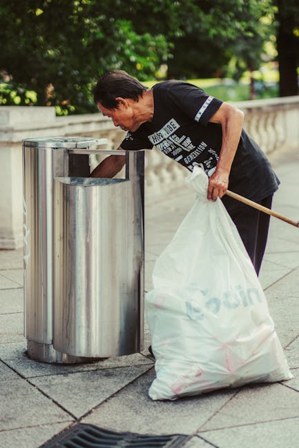 A middle-aged man with dark hair, wearing a black t-shirt with white printed text on the front, is seen disposing of rubbish into a tall, cylindrical stainless steel recycling bin located on a paved sidewalk. The man is bent forward slightly, holding a white plastic bag full of waste with one hand while using the other hand to insert waste into the opening of the bin. He appears to be engaged in a cleaning or waste collection activity in an outdoor urban environment. Behind him, there is a low stone or concrete wall with a decorative balustrade, and lush green trees and foliage in the background, providing a park-like setting. The scene is captured in natural daylight, suggesting a clear and bright day, emphasizing a neat and organized approach to rubbish disposal. The stainless steel bin has a reflective, polished surface, characteristic of modern waste management fixtures used in public spaces. This scene illustrates an example of private or independent rubbish handling, aligning with waste collection services available for community cleanup or street sanitation efforts.