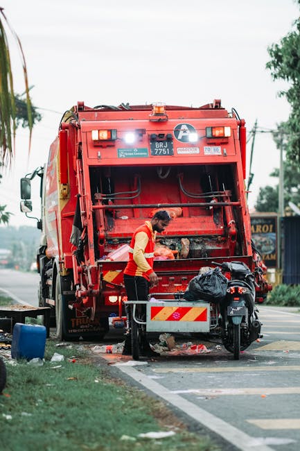 A worker wearing a yellow and red uniform is engaged in rubbish collection operation, standing beside the open rear loading compartment of a large red rubbish truck on a city street. The truck, with a white and red striped warning pattern on the back, is parked on the edge of the road near a grassy verge, with some discarded waste materials visible on the ground beneath the open bin. The vehicle's rear hydraulic mechanism is exposed, and a black trash bag is placed in the vicinity, suggesting it is recent or ongoing waste removal activity. In the background, there are power lines, trees, and a blue shopfront or container, indicating an urban environment. The scene is lit by natural daylight, with the sky overcast or partly cloudy, illustrating a routine street rubbish collection process typical of private waste management services such as House Clearance Finsbury Park, providing an example of on-site clearance and independent waste handling preparation.
