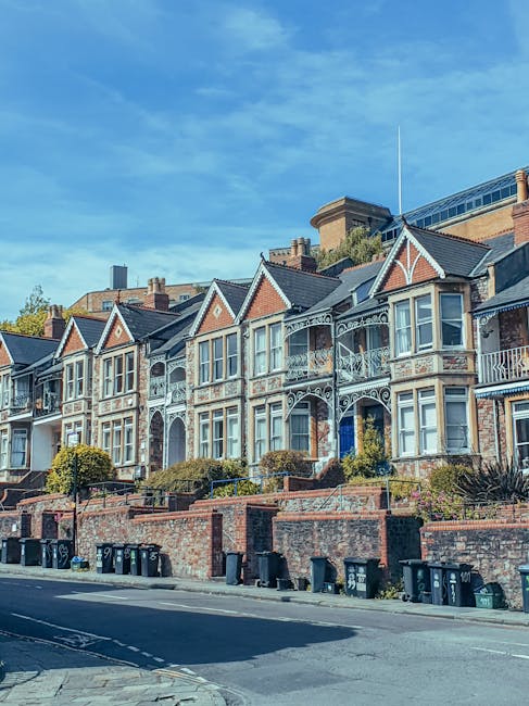 A street scene featuring a row of Victorian-style terraced houses with ornate wrought iron balconies and decorative gables, built with red and cream brickwork and large sash windows. The properties are elevated from the pavement, with brick retaining walls and small front gardens landscaped with shrubs and small trees. Several black wheelie bins are lined up along the pavement in front of the houses, indicating local waste disposal. The scene is illuminated by daylight, with a clear blue sky overhead and some modern buildings visible in the background. This setting suggests the importance of efficient rubbish collection and on-site disposal methods often employed by private waste management services like House Clearance Finsbury Park, especially for properties in densely populated urban areas.