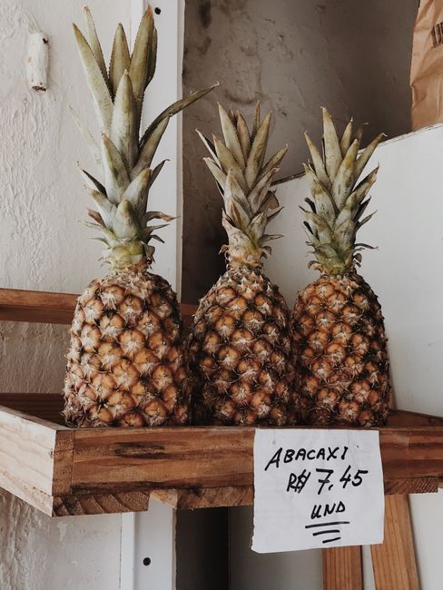 Three ripe pineapples with spiky green crowns sit upright in a wooden display crate on a shelf against a neutral-colored wall. The pineapples have textured, rough, and diamond-patterned brownish-yellow skin with some darker patches. The crowns are long, pointed, and slightly curved, with a mix of pale green and grayish tones. A handwritten sign attached to the crate indicates the price and type of fruit, with the word 'ABACAXI' and a price of 7.45, suggesting the pineapples are for sale at a market or shop. The background includes a portion of a textured wall and a faint glimpse of other elements, emphasizing a retail environment focused on fruit display, relevant for services involving the clearance or disposal of unwanted produce or similar items in a retail or market setting.