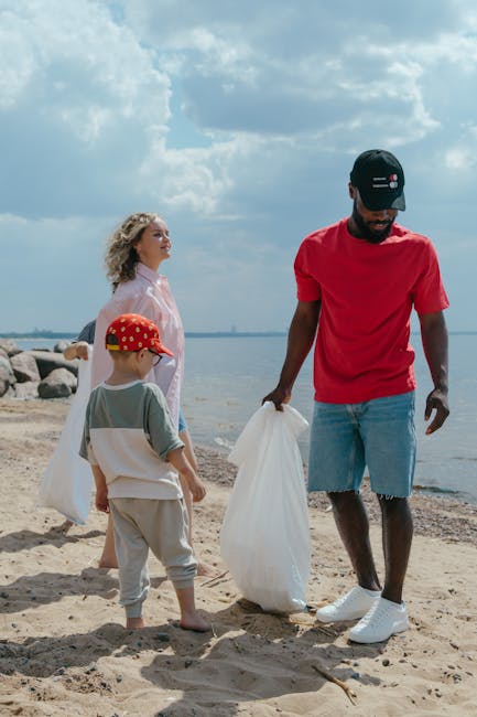 A man wearing a black cap, red t-shirt, and ripped denim shorts is standing on a sandy beach, holding a large white plastic bag filled with rubbish. Beside him, a young boy dressed in a light gray outfit with a red cap featuring yellow designs stands, also holding a white plastic bag, while a woman with curly hair, wearing a light pink blouse, looks on from behind. In the background, a rocky shoreline extends into a body of water under a partly cloudy sky, creating a natural setting that suggests a beach clean-up activity, consistent with private waste handling or rubbish removal services like those offered by House Clearance Finsbury Park.