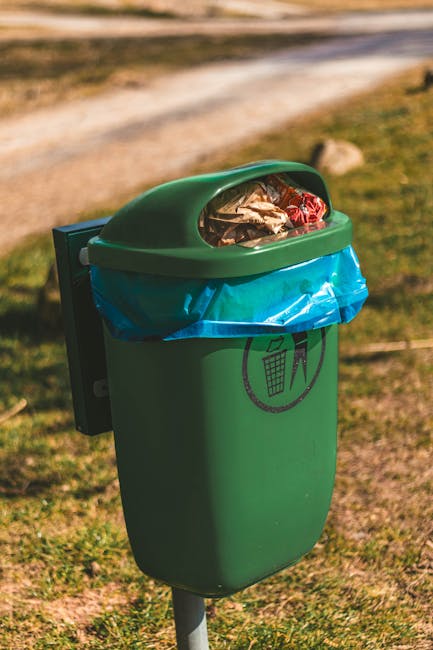 A green outdoor rubbish bin with a rounded top and a semi-transparent lid, filled with mixed waste including paper, plastic bottles, and foil wrappers, situated on a metal post in a grassy area with patches of dirt and a gravel pathway in the background. The bin has a black recycling symbol with a trash can icon printed on its front, indicating its use for general waste collection. The environment appears to be a park or open public space with natural sunlight illuminating the scene; the setting suggests an emphasis on proper waste disposal, aligning with private waste handling services offered by House Clearance Finsbury Park for rubbish removal and site clearances.