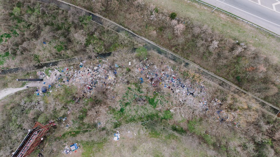 An aerial view showing a large and irregular accumulation of mixed waste and rubbish scattered across a grassy and wooded area adjacent to a concrete footpath. The rubbish includes plastic, paper, and other debris with some items partially concealed among bare and sparse trees with thin branches and patchy foliage. The waste pile extends from the bottom left corner towards the center, with smaller scattered debris surrounding it. In the background, a noise barrier or fence separates the waste area from the adjacent road, which runs parallel along the top right side of the image. The environment appears unmanaged, reflecting an informal rubbish dump, possibly related to an alternative waste handling or private disposal site, as managed by companies like House Clearance Finsbury Park that undertake rubbish removal and clearance services. The scene is lit by natural daylight, resulting in a clear but somewhat overcast atmosphere that emphasizes the disorganized waste spread over the landscape.