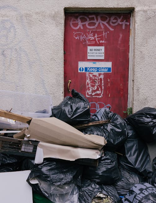 A large pile of black rubbish bags, some partially torn or bulging, stacked in front of a red metal door set into a plain, off-white textured building wall. The bags contain waste materials and are placed against the wall, with a few cardboard boxes and loose debris on top of or among them. The door features various graffiti tags in white and pink spray paint, along with a small white sign stating 'NO MONEY' and another blue and white sign reading 'Keep clear.' The scene suggests a setting awaiting waste removal or on-site clearance, typical of private rubbish disposal services. The lighting appears natural, illuminating the scene evenly without harsh shadows, emphasizing the contrast between the packed rubbish and the plain exterior wall. This image reflects an environment where independent waste collection or rubbish clearance from premises like flats or small commercial buildings might take place, aligning with services offered by companies such as House Clearance Finsbury Park for urgent or same-day rubbish removal needs.
