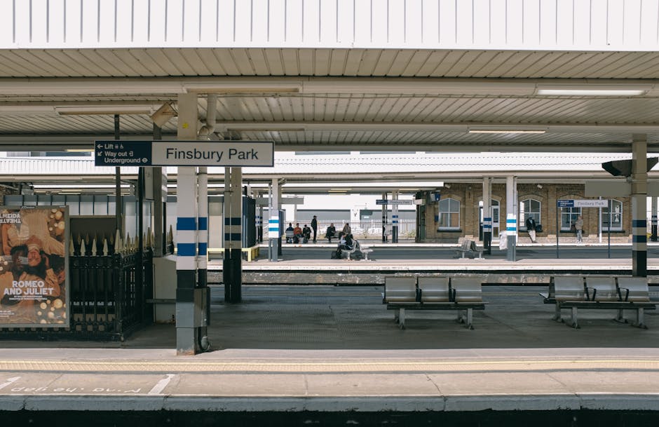 View of Finsbury Park train station platform with a central sign displaying the station name, surrounded by a covered waiting area. The platform features several metal benches with wooden slats, positioned on a paved surface. In the background, there are additional platforms, railway tracks, and a few passengers waiting or walking. On the left side, a poster advertising a theatrical performance is visible on a black metal fence. The station structure includes concrete supports and a metal roof with a light-colored finish, creating an outdoor yet sheltered environment. The lighting appears natural, suggesting daytime, and the scene is clean and orderly, typical of urban railway stations. This environment reflects the standard context in which private or independent rubbish removal services may operate, such as clearing waiting areas or station surroundings associated with urban transport infrastructures. The scene demonstrates a typical public transport hub that could be part of or adjacent to a collection or clearance operation managed by professional waste management providers like House Clearance Finsbury Park.